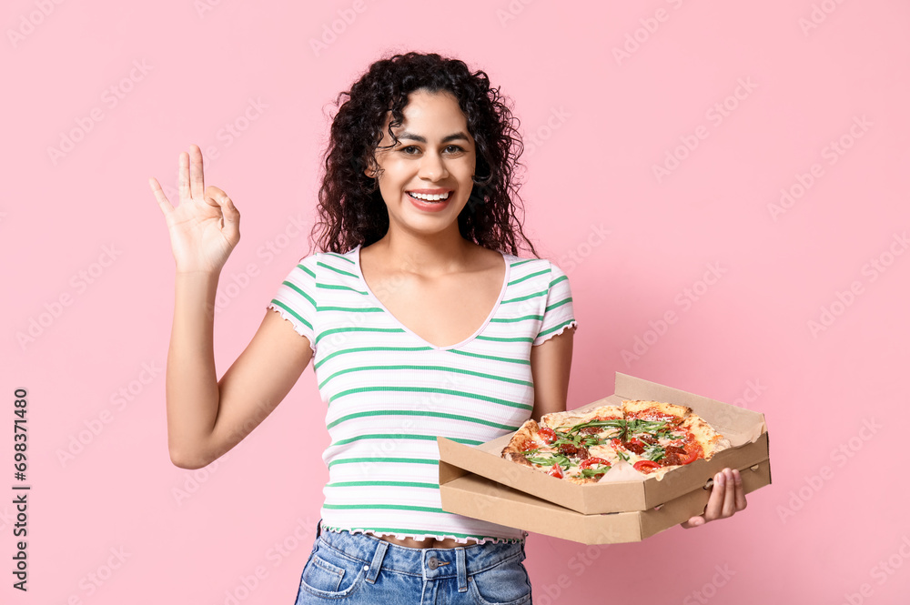 Beautiful young happy African-American woman with delicious pizza showing OK gesture on pink background