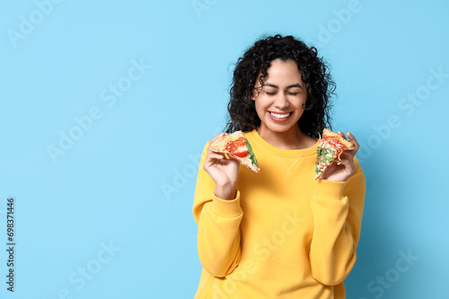 Beautiful young happy African-American woman with slices of delicious pizza on blue background