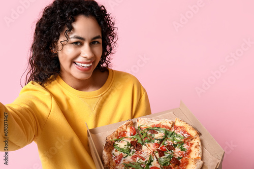 Beautiful young happy African-American woman with delicious pizza taking selfie on pink background