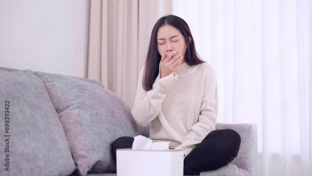 Asian woman coughing and sneezing, feeling anxious about getting sick or unwell while sitting on her home sofa. Capturing the moment of discomfort and concern at home Asian woman coughing and sneezing, feeling anxious about getting sick or unwell while sitting on her home sofa. Capturing the moment of discomfort and concern at home