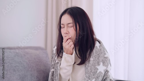 Asian woman coughing and sneezing, feeling anxious about getting sick or unwell while sitting on her home sofa. Capturing the moment of discomfort and concern at home photo
