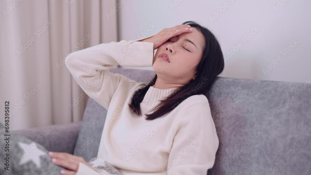 Asian woman feeling a headache and body warmth, anxious about getting sick or unwell while sitting on her home sofa and wrapped in a blanket. Capturing the moment of discomfort and concern at home Asian woman feeling a headache and body warmth, anxious about getting sick or unwell while sitting on her home sofa and wrapped in a blanket. Capturing the moment of discomfort and concern at home