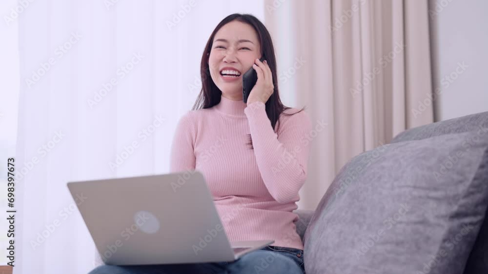 Asian woman happily chatting on the phone and working on her laptop while sitting on the sofa at home. A positive and productive remote work environment. Asian woman happily chatting on the phone and working on her laptop while sitting on the sofa at home. A positive and productive remote work environment.