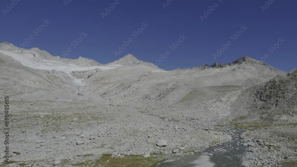 Drone captures a man approaching a small bridge on a glacier river trail. The journey continues to showcase the Möseler-Mesule and the almost completely melted and Neves-Nofesferner glacier.