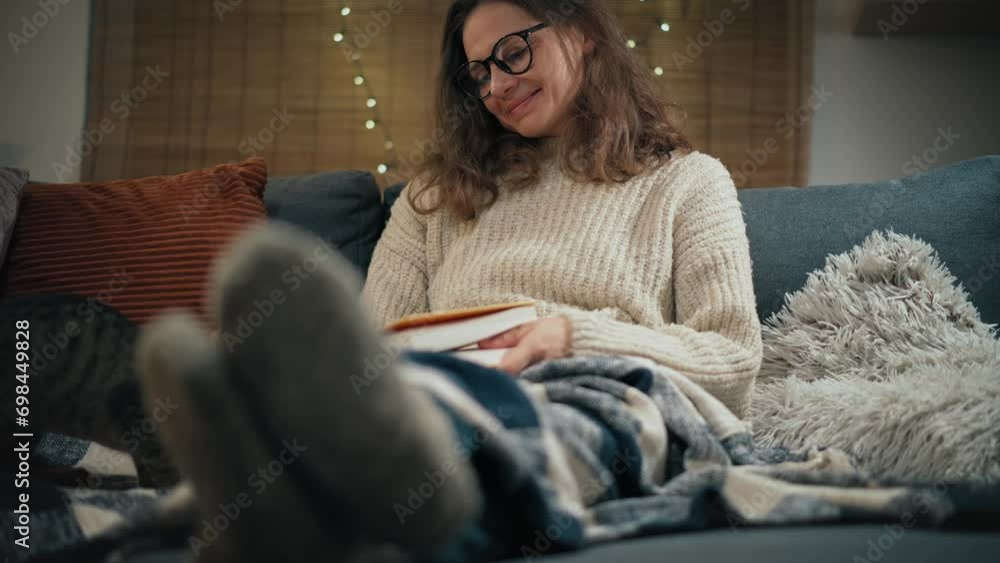 A young Caucasian woman reading a book sitting on the couch with her cute grey cat at home on a winter evening A young Caucasian woman reading a book sitting on the couch with her cute grey cat at home on a winter evening