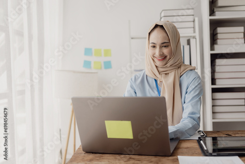 Photography Young Asian Muslim woman in simple clothes working with laptop computer in modern business office on documentation and analysis project, energetic digital entrepreneur working on startup project