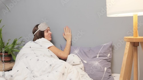 Unhappy young woman in sleeping mask on her head suffering and disturbed by noisy neighbors calling on smartphone knocking to wall trying to falling sleep in bed at home at night. photo