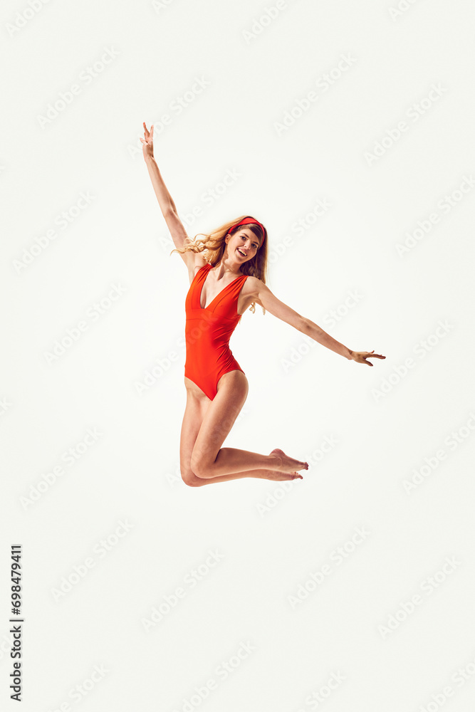 Beautiful young blonde woman in stylish red vintage swimsuit cheerfully jumping against white background. Concept of summer vacation, travelling, retro style, fashion, relaxation, emotions