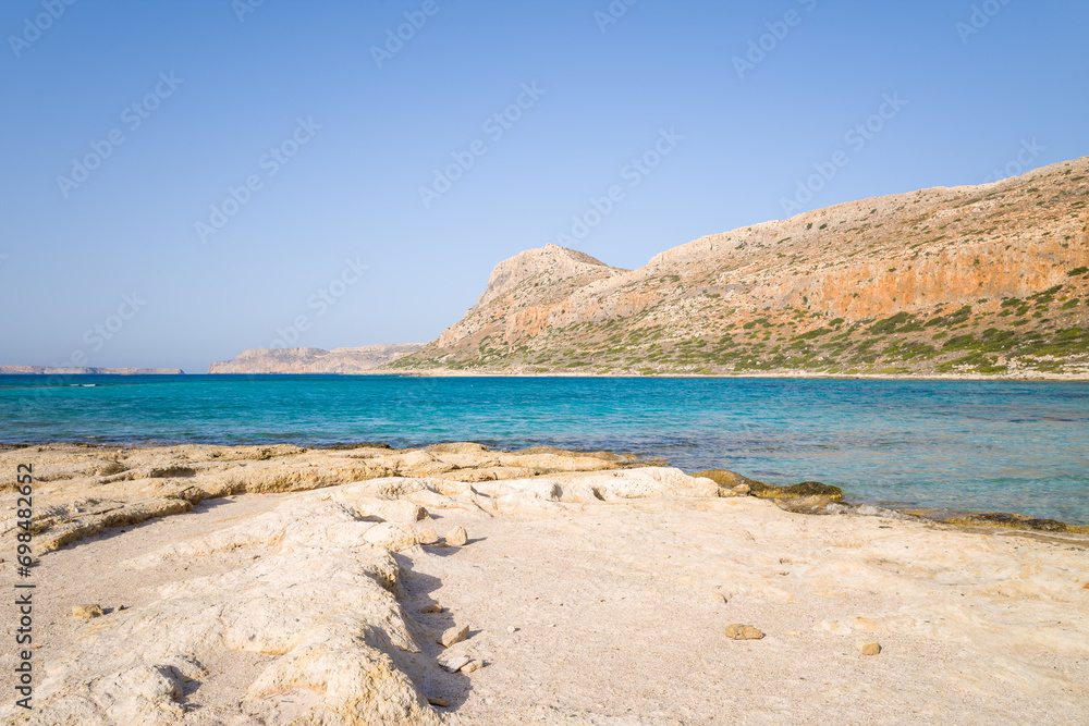 The sandy beach with pink reflections at the foot of the rocky cliffs, in Europe, Greece, Crete, Balos, By the Mediterranean Sea, in summer, on a sunny day. The sandy beach with pink reflections at the foot of the rocky cliffs, in Europe, Greece, Crete, Balos, By the Mediterranean Sea, in summer, on a sunny day.