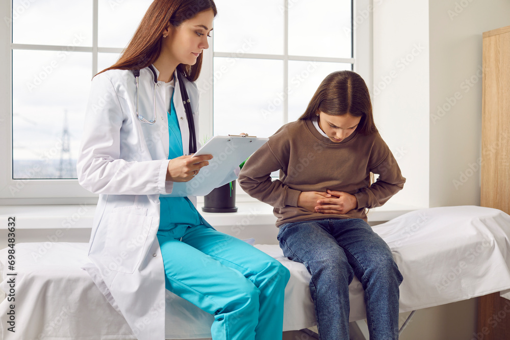 Teenage girl sitting on the couch in the doctor's office and pointing to her stomach to paediatrician during medical examination in clinic. Child doctor listening to the patient's complaints.