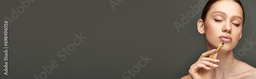 portrait of young woman holding syringe near face on grey background, lip enhancement banner photo