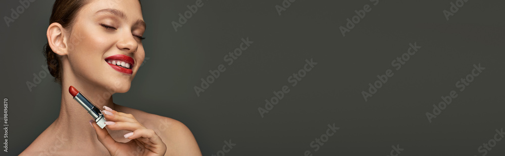 cheerful young woman with red lips holding lipstick and smiling on grey background, makeup banner cheerful young woman with red lips holding lipstick and smiling on grey background, makeup banner