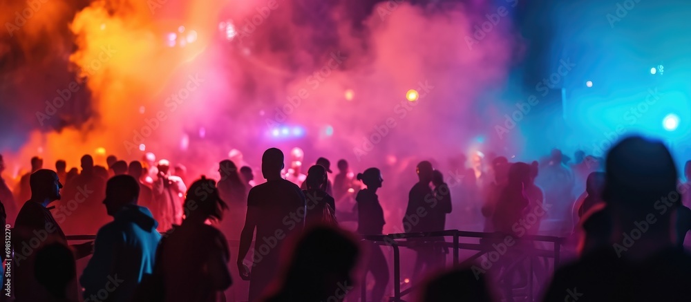 Multicolored lights, smoke, and silhouettes of people on stage create a blurry background for design. Multicolored lights, smoke, and silhouettes of people on stage create a blurry background for design.