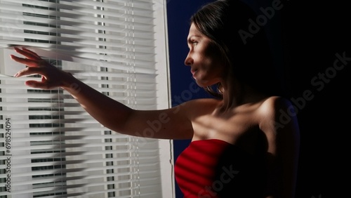 Portrait of female model in the studio. Appealing woman in red dress close up shot standing near window opens looks out through jalousie.