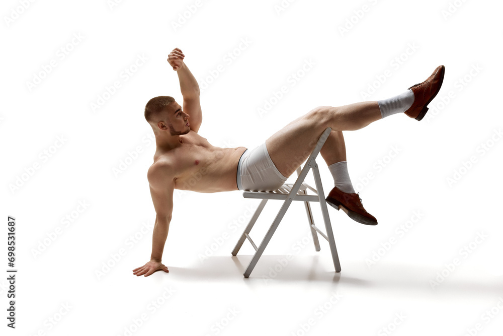 Young man, shirtless model with perfect body shapes sits posing on chair with her legs hanging over back of it against white studio background. Concept of men's health, beauty, fitness. Body art Young man, shirtless model with perfect body shapes sits posing on chair with her legs hanging over back of it against white studio background. Concept of men's health, beauty, fitness. Body art