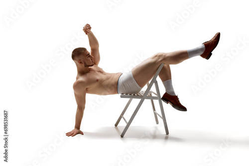 Young man, shirtless model with perfect body shapes sits posing on chair with her legs hanging over back of it against white studio background. Concept of men's health, beauty, fitness. Body art © Lustre Art Group 