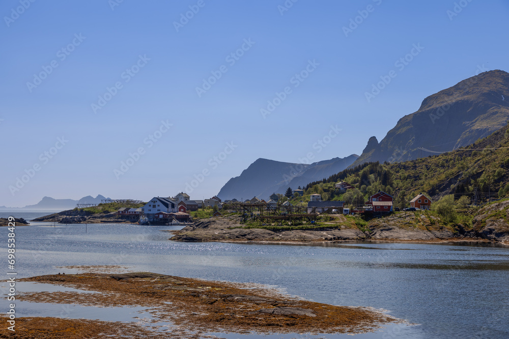 Moskenes in the Lofoten Islands, Norway, gleams under a sunny, blue sky, featuring traditional cod drying racks near houses, set against a backdrop of green mountains