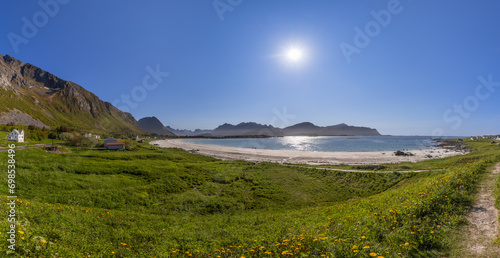 Wide-angle panorama showcasing the radiant sun over Jusnesvika Bay, with lush greenery, yellow wildflowers, white sandy Rambergstranda beach, and people basking in the sun photo