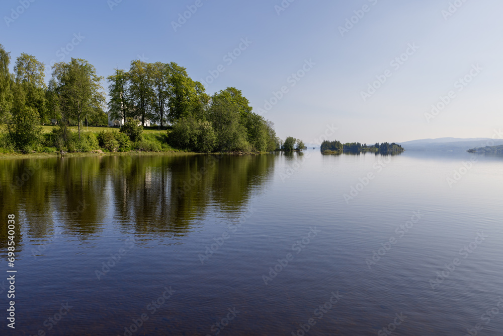 The serene Lake Snasa in Steinkjer, Norway, mirrors a wooden Kvam Church, nestled among dense trees along its tranquil shores