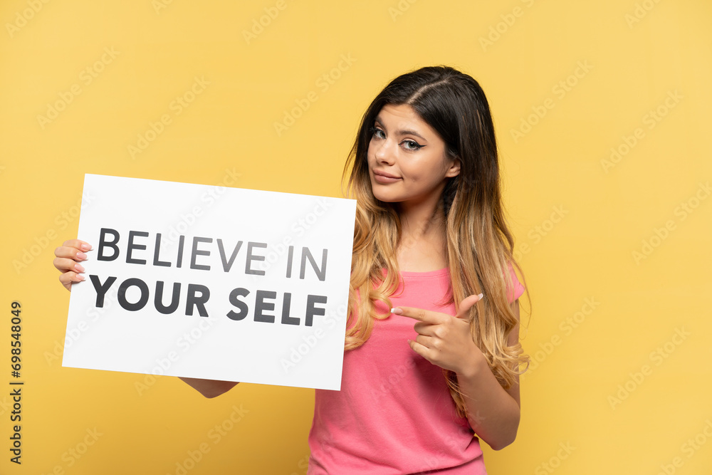 Young Russian girl isolated on yellow background holding a placard with text Believe In Your Self and pointing it Young Russian girl isolated on yellow background holding a placard with text Believe In Your Self and pointing it