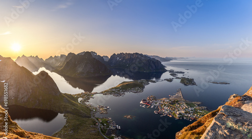 The sun hangs low in the expansive sky over Reinebringen, offering a super panorama of Lofoten Islands. View of Reine's vibrant fishing village amidst the tranquil waters and rugged mountain terrain photo