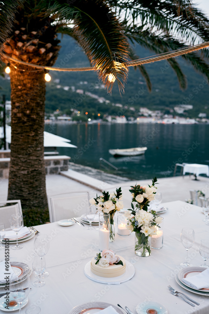 Wedding cake stands on a plate on a table near a palm tree with luminous garlands on the pier