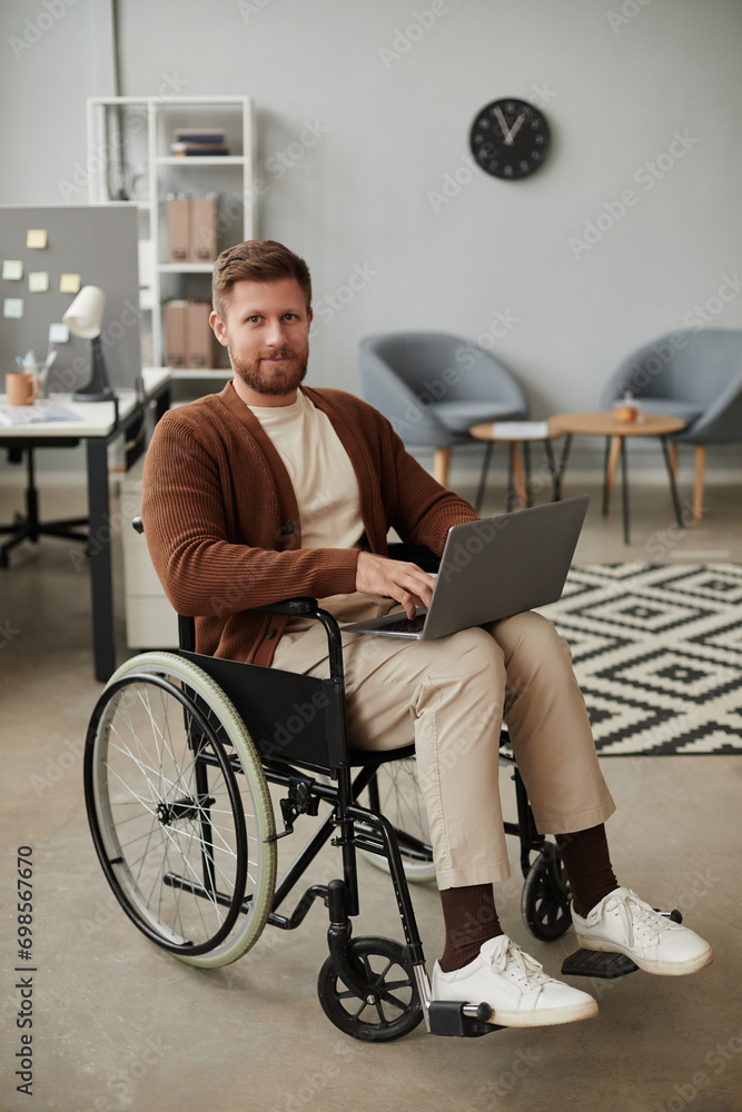 Vertical full length portrait of bearded man with disability using laptop in wheelchair and looking at camera Vertical full length portrait of bearded man with disability using laptop in wheelchair and looking at camera