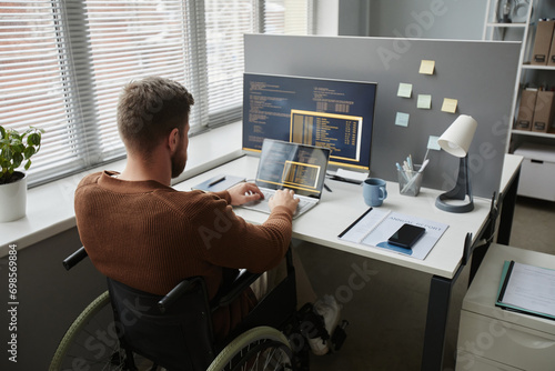 Back view of adult man with disability using wheelchair at office workplace and coding in IT development