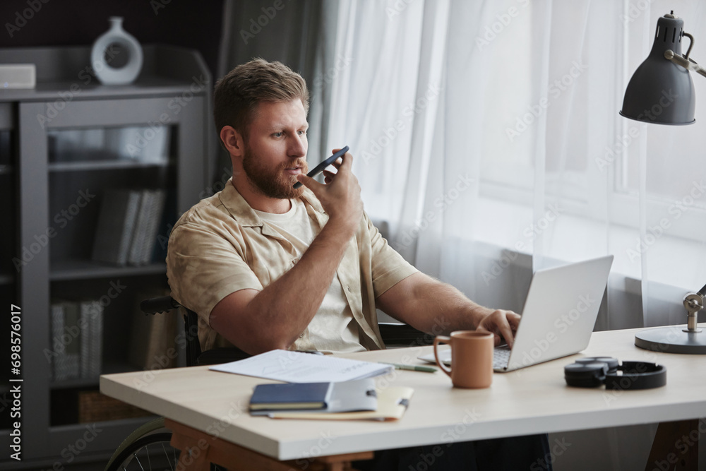 Portrait of bearded adult man with disability working at home office workplace and recording voice message via smartphone, copy space
