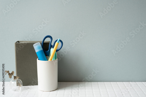 Notebook and holder with glue stick, pen, scissors on white tile desk. gray wall background