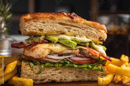 Obraz na płótnie Grilled chicken breast sandwich with lettuce, tomato, bacon, red onion rings and avocado served on artisan bread and a portion of French fries w salonie