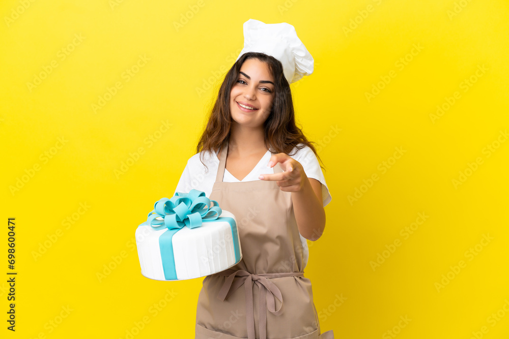 Young caucasian pastry chef woman with a big cake isolated on yellow background pointing front with happy expression Young caucasian pastry chef woman with a big cake isolated on yellow background pointing front with happy expression
