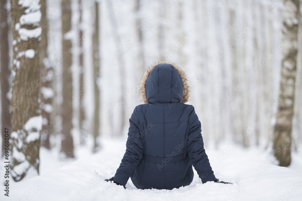 Young adult woman sitting on fresh white snow at birch tree forest. Looking far away. Spending time alone in beautiful cold winter day. Back view. Thinking about life. Peaceful atmosphere in nature. Young adult woman sitting on fresh white snow at birch tree forest. Looking far away. Spending time alone in beautiful cold winter day. Back view. Thinking about life. Peaceful atmosphere in nature.