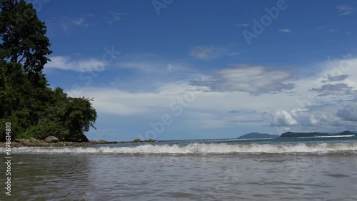 The ocean waves roll towards the shore, foaming and spreading on the beach. Green vegetation against a background of blue sky and clouds. Islands in the distance. Malaysia. Borneo. Kota Kinabalu.