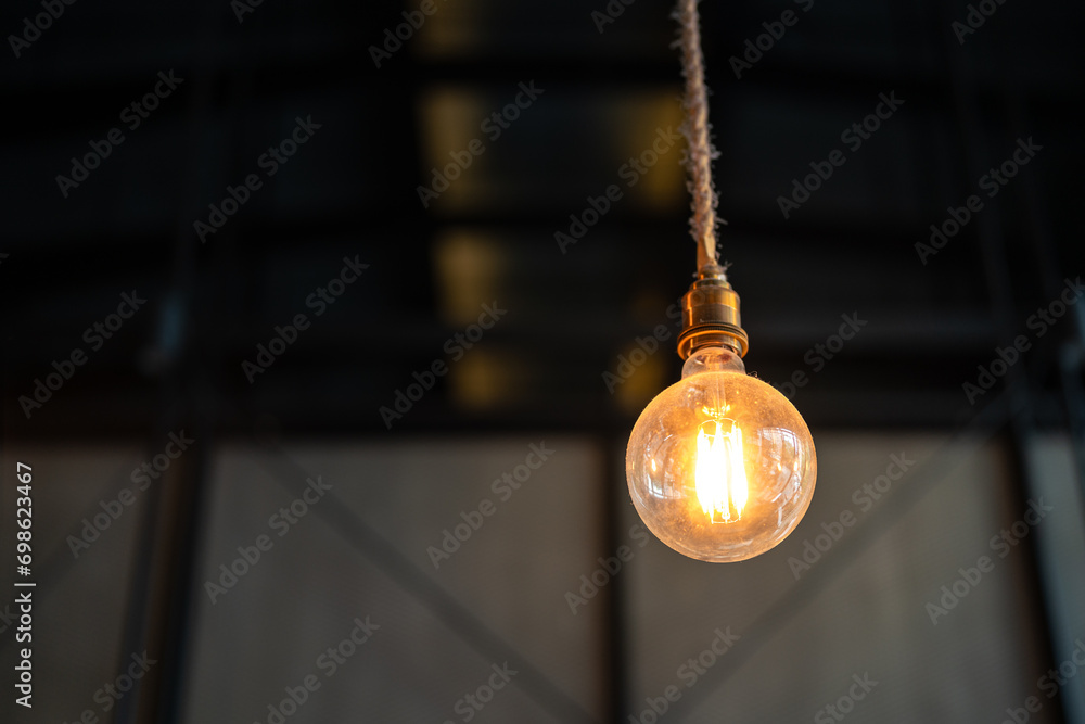 A classic style lightbulb is glowing in orange warn lighting is hanging down from ceiling with rope. Interior decoration equipment object photo. Close-up and selective focus.