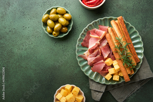 Slices of prosciutto or jamon. Delicious grissini sticks with prosciutto, cheese, rosemary, olives on green plate on dark background. Appetizers table with italian antipasto snacks Top view copy space photo