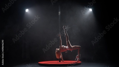 Portrait of young female dancer in the studio. Professional pole dancer girl spinning on pylon, showing modern pole performance, black background with smoke.