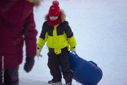 A little boy in a warm yellow jacket carries a sled up the hill to slide down