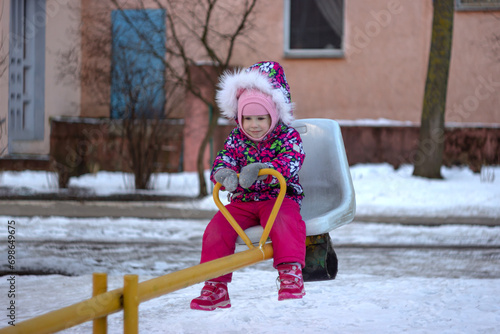 A little girl in a warm pink winter jacket rides on a swing at the playground