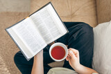 Close up, young woman drinking hot tea and reading favorite paper book, top view.