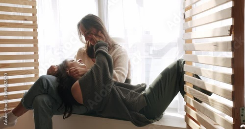 Two lesbian woman relaxing in cuddling on window sill, talking and touching each other tender. Female couple in romantic relationship enjoy spend time together. Same sex love partners photo