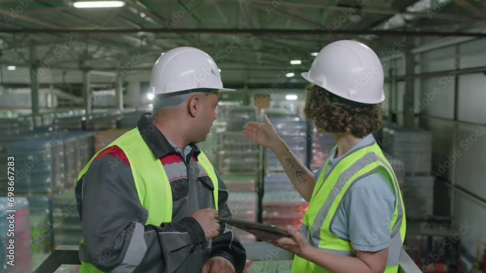 Medium shot of two workers discussing delivery document using digital tablet at warehouse with pallets in background Medium shot of two workers discussing delivery document using digital tablet at warehouse with pallets in background