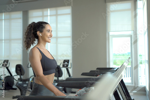 Happy healthy hispanic woman practicing a cardio exercise by running and walking on treadmill machine in fitness and using fitness tracker or smartwatch to track workout progress.