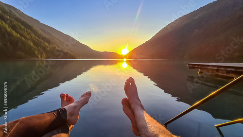 Couple enjoying allure of sunset at alpine lake Weissensee in remote Austrian Alps in Carinthia. Hanging legs of people. Embrace serene ambiance as tranquil surface of water amplifies sun gentle rays photo