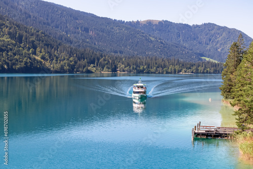 Passenger ferry boat gliding along east bank of alpine lake Weissensee, Carinthia, Austria. Glistening turquoise water mirrors the beauty of untouched nature. Scenic view of tranquil forest in summer