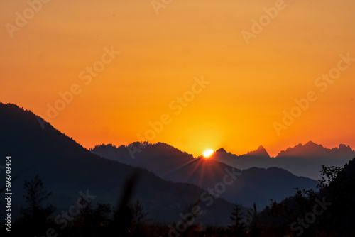 Silhouette of majestic mountain peaks against backdrop of vibrant sunset. Panoramic viewing point at east bank of Weissensee in Carinthia, Austria. Gazing through tranquil forest, romantic atmosphere