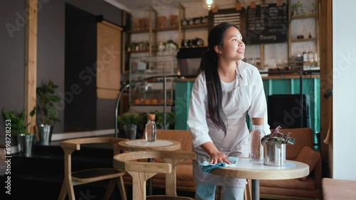 Smiling asian woman waitress in apron wiping tables in cafe photo