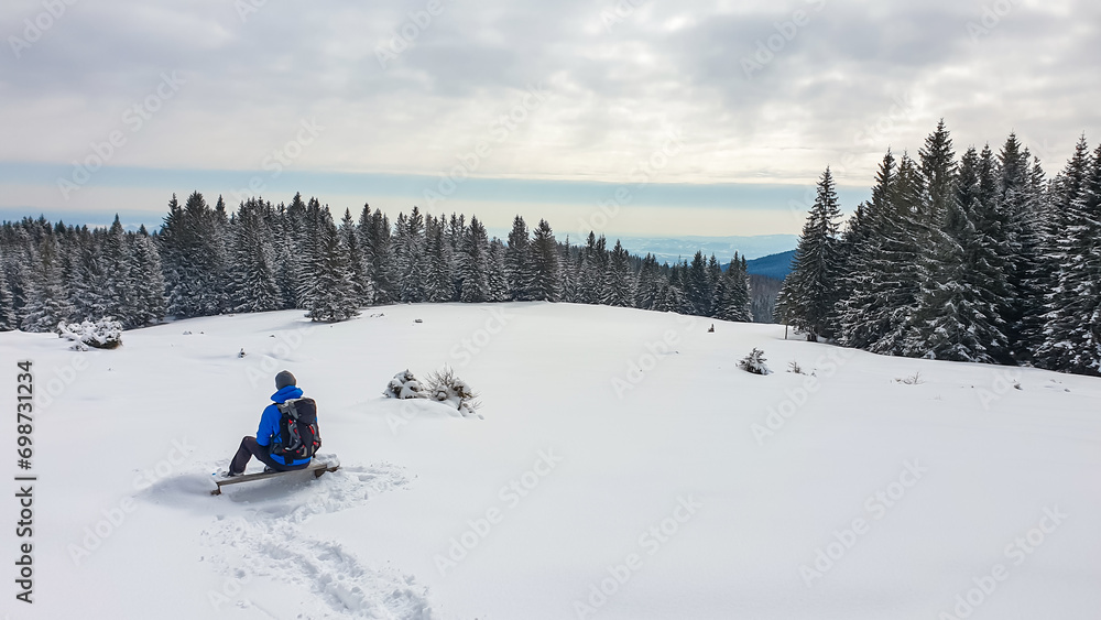 Man sitting on alpine snowy meadow with panoramic view of mystical forest on Reinischkogel, Koralpe, Lavanttal Alps, Styria, Austria. Winter wonderland with snow covered tall pine trees, Austrian Alps