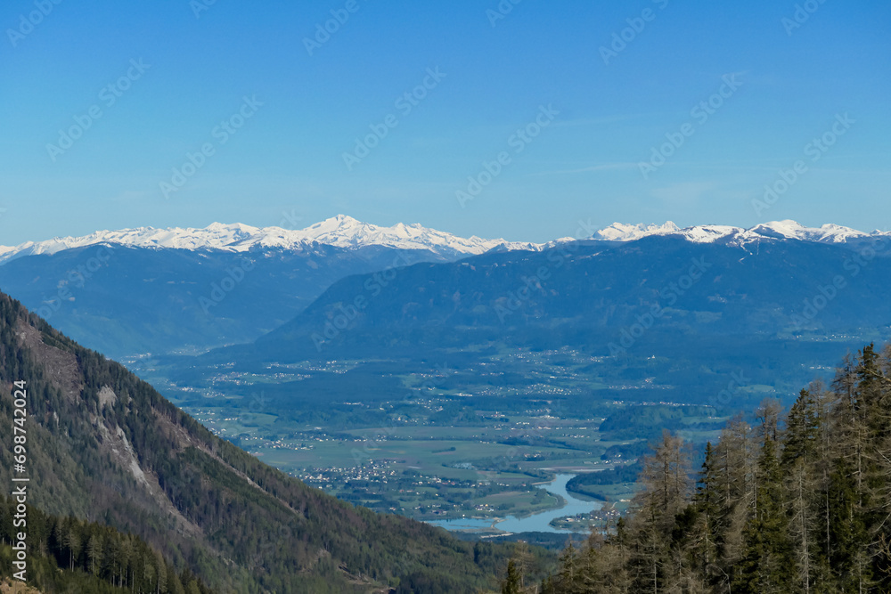 Scenic view on Sinacher Gupf in Karawanks mountains and the Drava river in Rosental valley in Carinthia, Austria. Forest in early spring. Snow capped Hohe Tauern mountain range can be seen. Sunny day