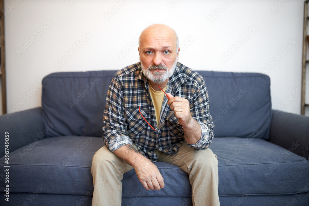 Indoor portrait of worried anxious grandfather in plaid shirt having rest sitting on couch in living room holding glasses, thinking of his life, world problems, health and aging process Indoor portrait of worried anxious grandfather in plaid shirt having rest sitting on couch in living room holding glasses, thinking of his life, world problems, health and aging process